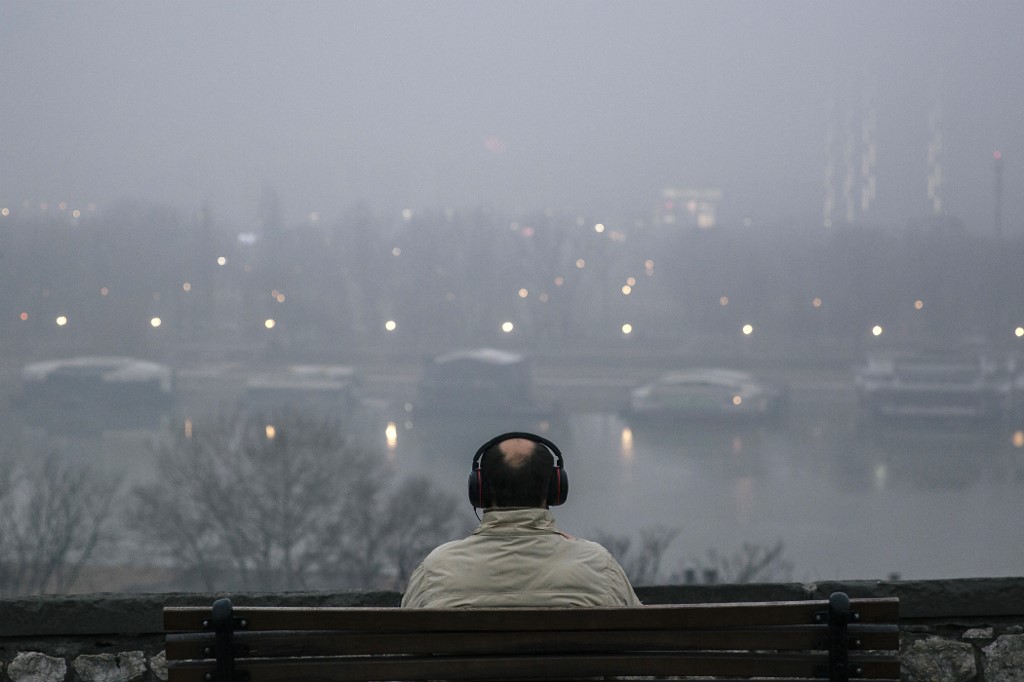 A man wearing headphones sits on a bench and looks towards the Sava river, as heavy fog and air pollution dominate the sky over Serbian capital Belgrade on January 13, 2020. - A thick cloud of pollution has been visible over the city for several days now, and according to the AirVisual app, Belgrade has several times in the last week been among the most polluted cities in the world. (Photo by Vladimir Zivojinovic / AFP)